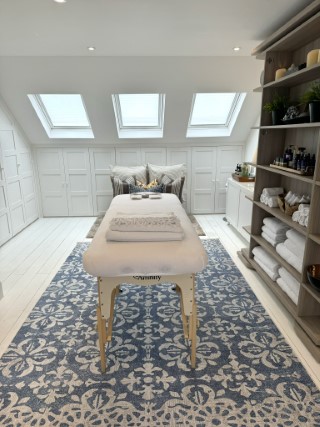 A massage table, with a clean white towelling cover.  The table is on a large blue and white rug. A blue ottoman in the background.